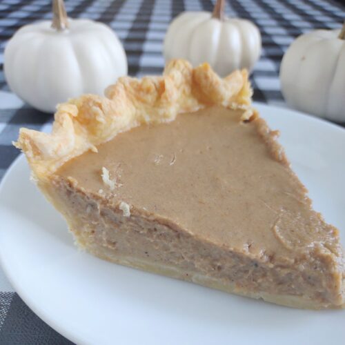 Creamy homemade pumpkin pie with flaky crust on a white plate, autumn decor with mini white pumpkins in background.