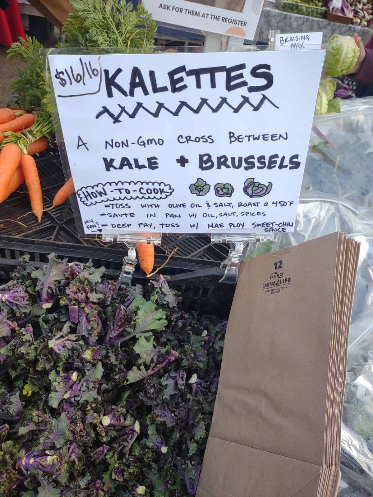A sign for Kalettes - a cross between Kale and Brussels Sprouts in front of a container filled with Kalettes at a farmers market.