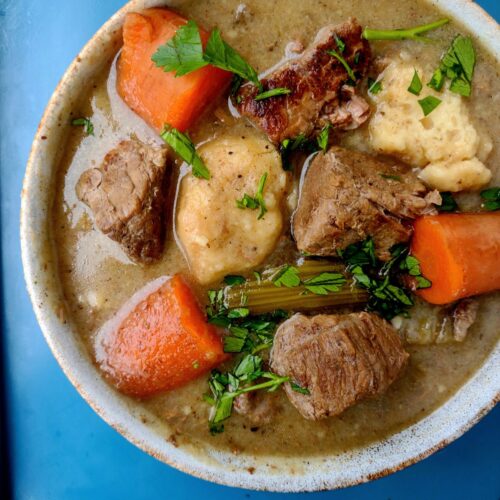 A bowl filled with beef stew meat, carrots, celery, and dumplings sitting on a blue sheet pan