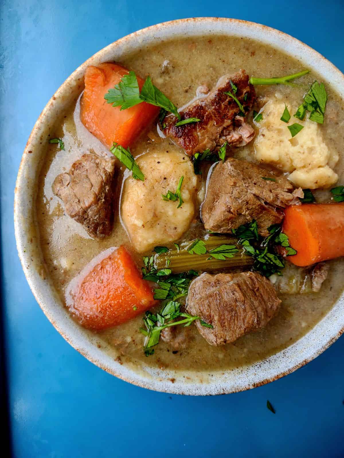 A bowl filled with beef stew meat, carrots, celery, and dumplings sitting on a blue sheet pan