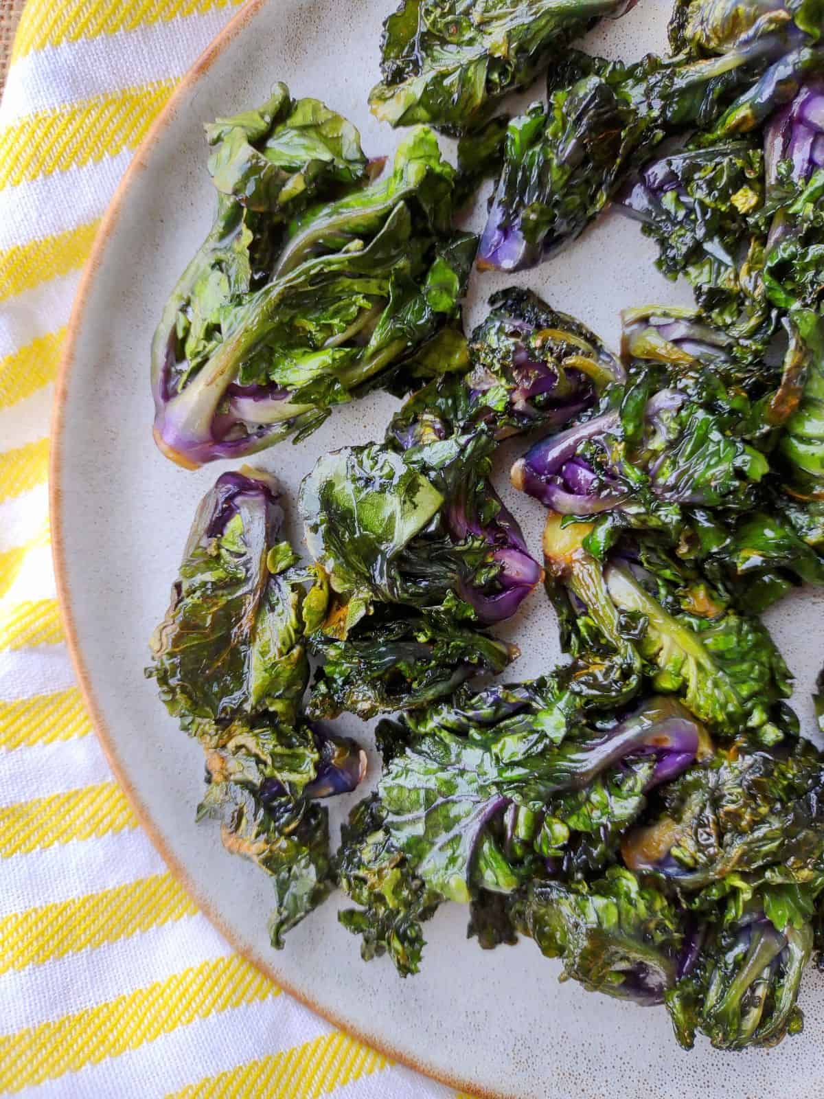 Sautéed green and purple Kalettes on a plate with a yellow & white striped towel underneath.