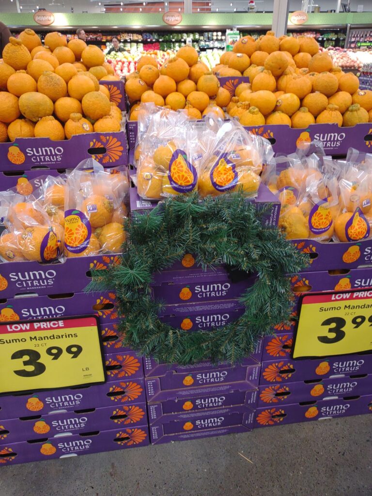 A display of Sumo Citrus mandarins at a Fred Meyer store near Christmas time. The fruit is on purple boxes loose and in bags with a Christmas wreath attached to the boxes.