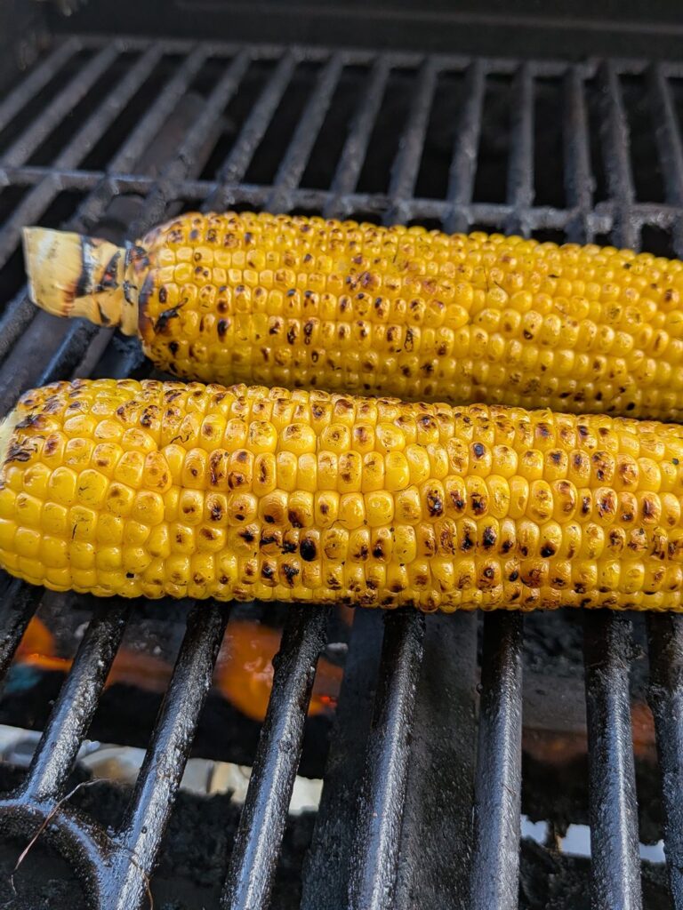 Two yellow ears of corn on the grill with char marks on them. The grill is on as a flame can be seen.