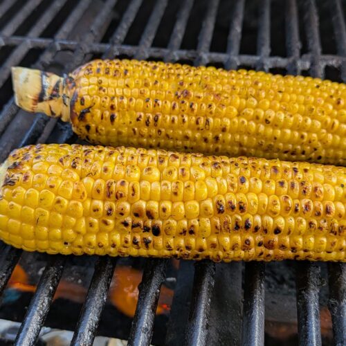 Two yellow ears of corn on the grill with char marks on them. The grill is on as a flame can be seen.