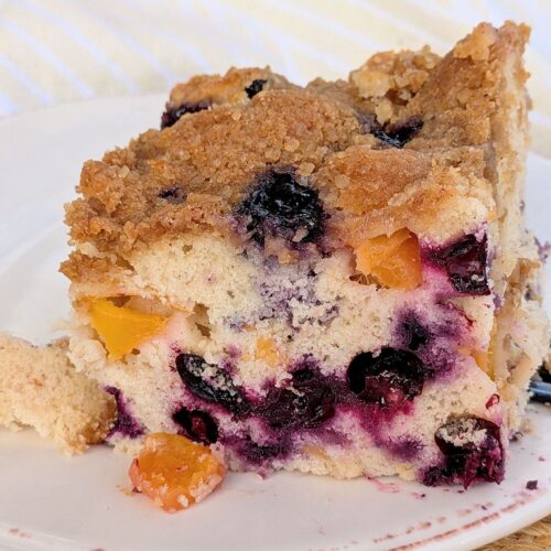 A side view of a slice of Blueberry-Peach buckle showing the fruit inside the cat. The buckle is on a white plate with a brown accent.