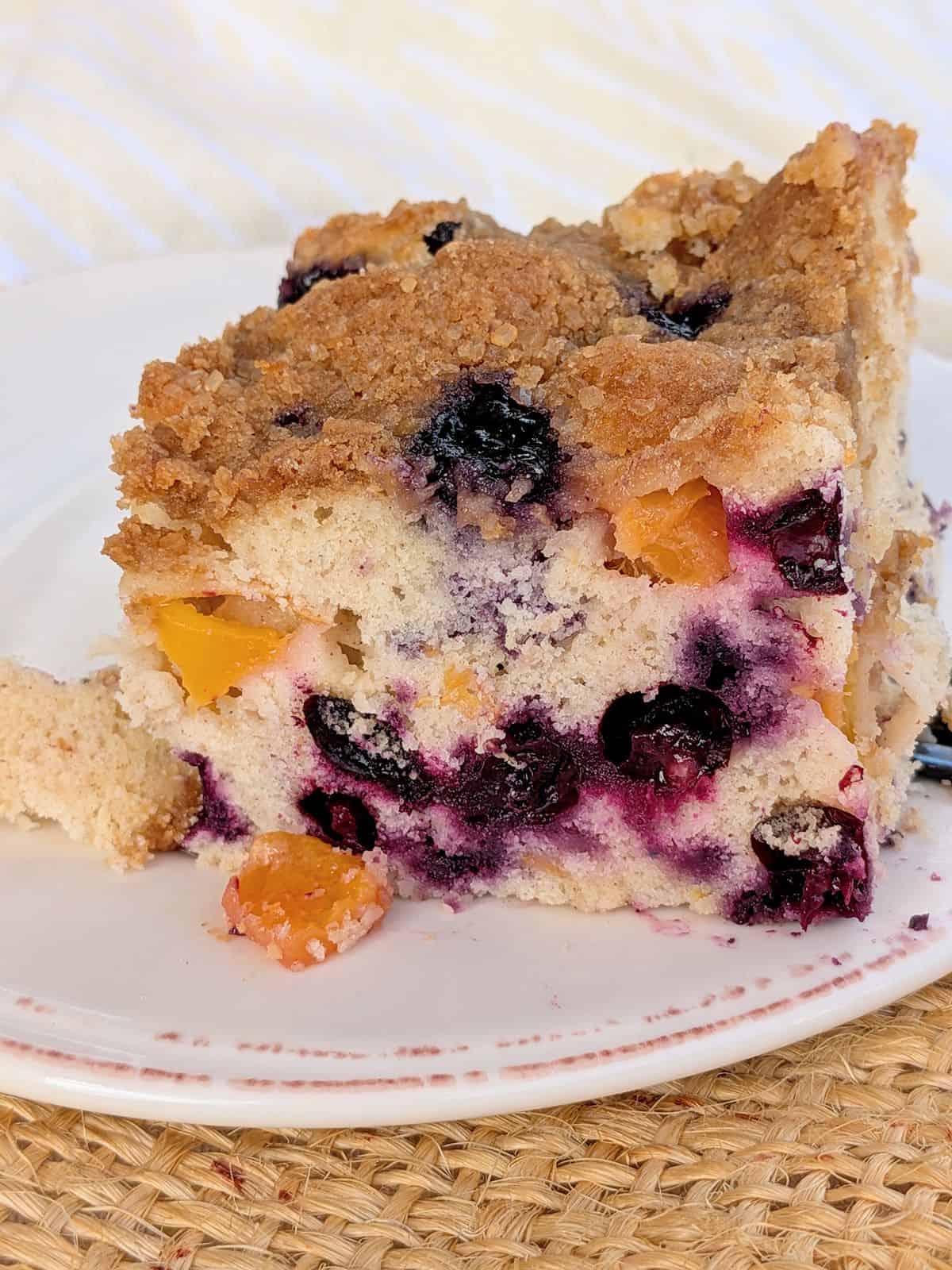 A side view of a slice of Blueberry-Peach buckle showing the fruit inside the cat. The buckle is on a white plate with a brown accent.