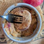A single serving air fryer apple cobbler in a small round cake pan with a black fork.