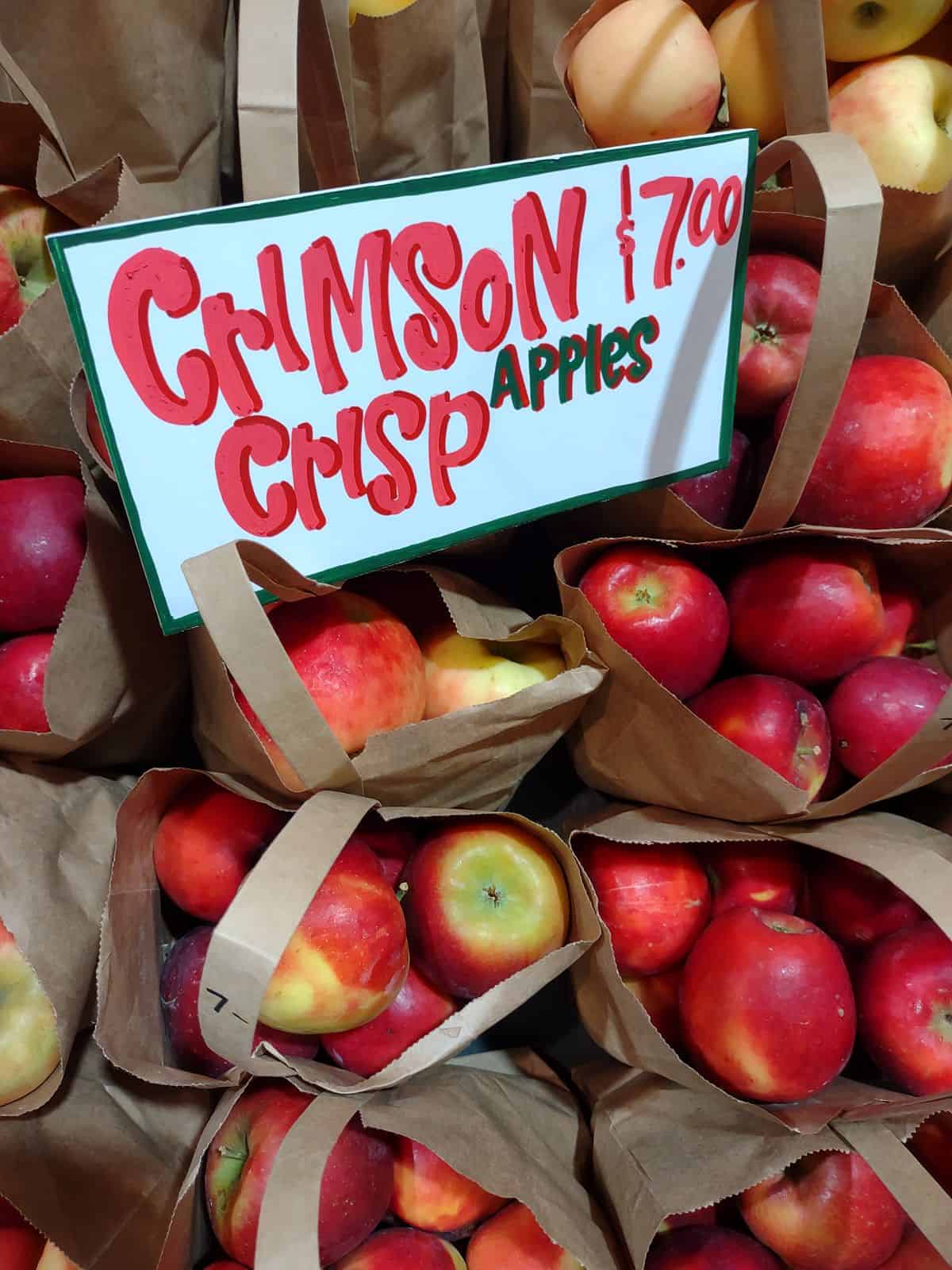 Paper bags of Crimson Crisp apples on a market with a sign that says they are $7.00 for a bag.