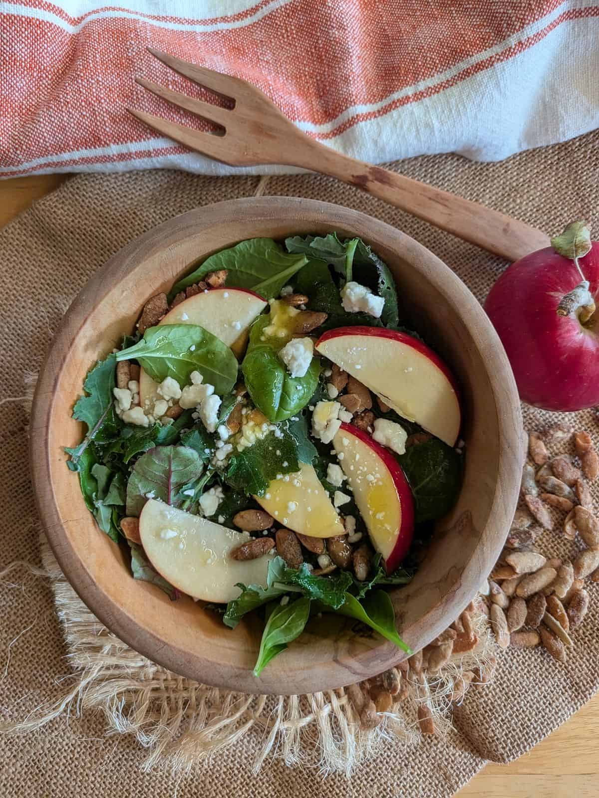 A kale and chard salad with sliced Sweet Sixteen apple, pumpkin seeds, and feta cheese in a wood bowl.